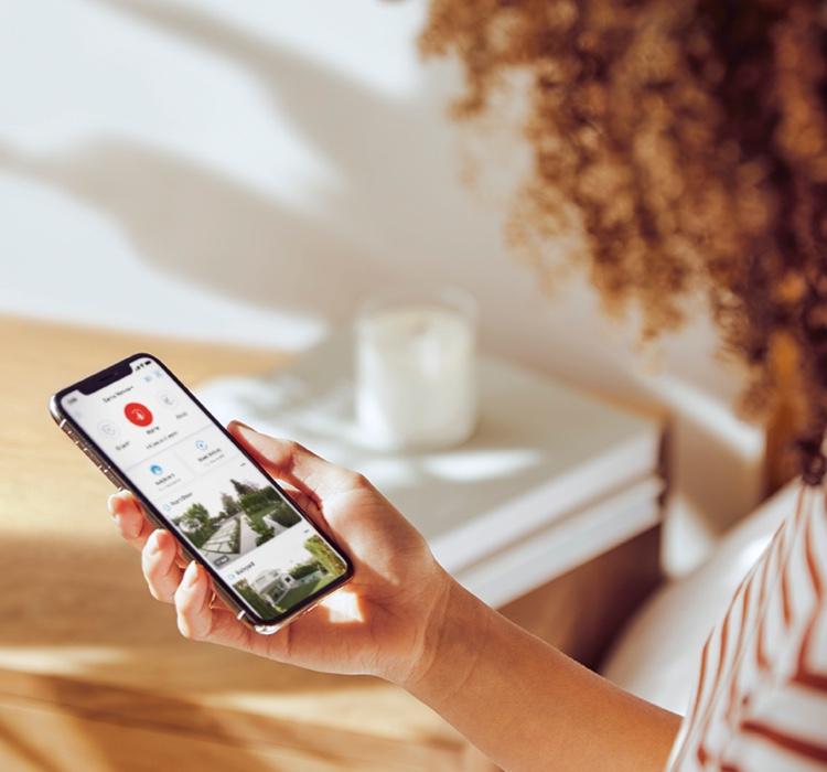 Woman holding a smartphone displaying the Ring App, monitoring her home security feed in a well-lit room.