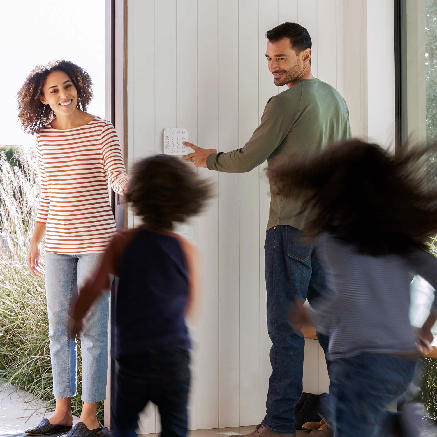 Protected Enhanced Kit (Battery Doorbell Plus + Alarm Security Kit, 8-Piece) - Smiling woman greets two children running toward her, while a man arms a Ring security keypad near the open door.