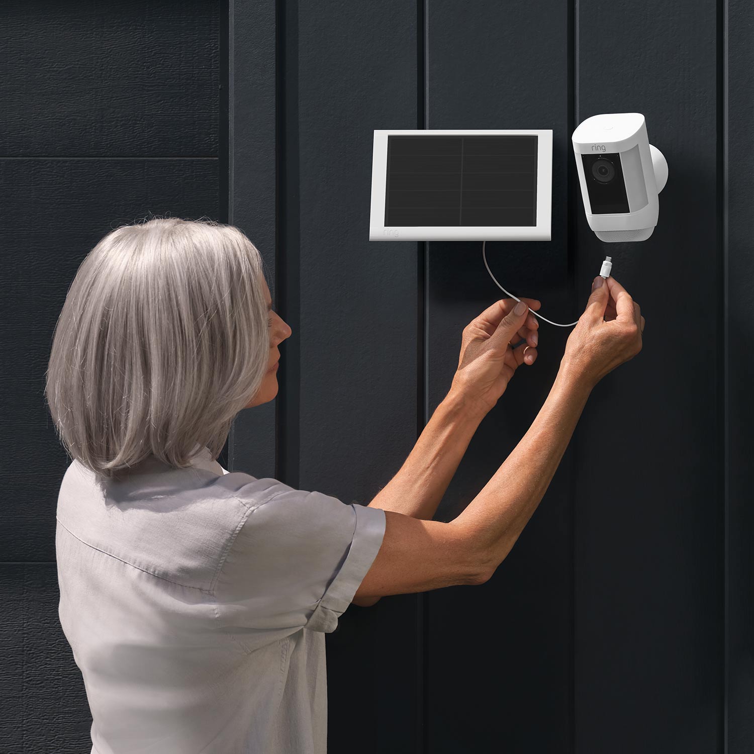 Spotlight Cam Pro (Featuring Retinal 2K) - Solar:Woman connecting a white Ring Solar Panel to a white Spotlight Cam Pro mounted on a dark exterior wall.