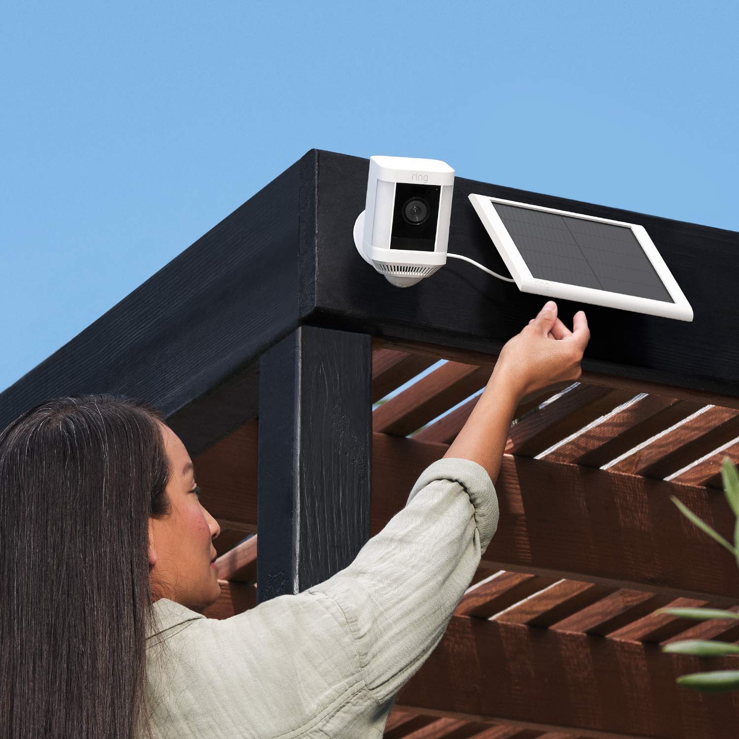 Spotlight Cam Plus - Solar:Woman adjusting Ring Solar Panel and Ring Spotlight Cam Plus, both mounted on dark wooden patio beam under blue sky.